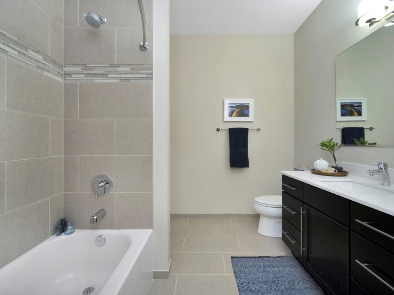 This image shows a contemporary bath that has quartz countertops, ceramic tile floors, and tub surrounds. It has an aisle directly passing to the kitchen area.
